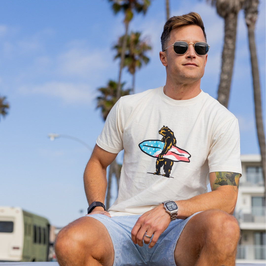Man wearing a white t-shirt with a bear holding a surfboard and American flag design, sitting on a beach with palm trees in the background.
