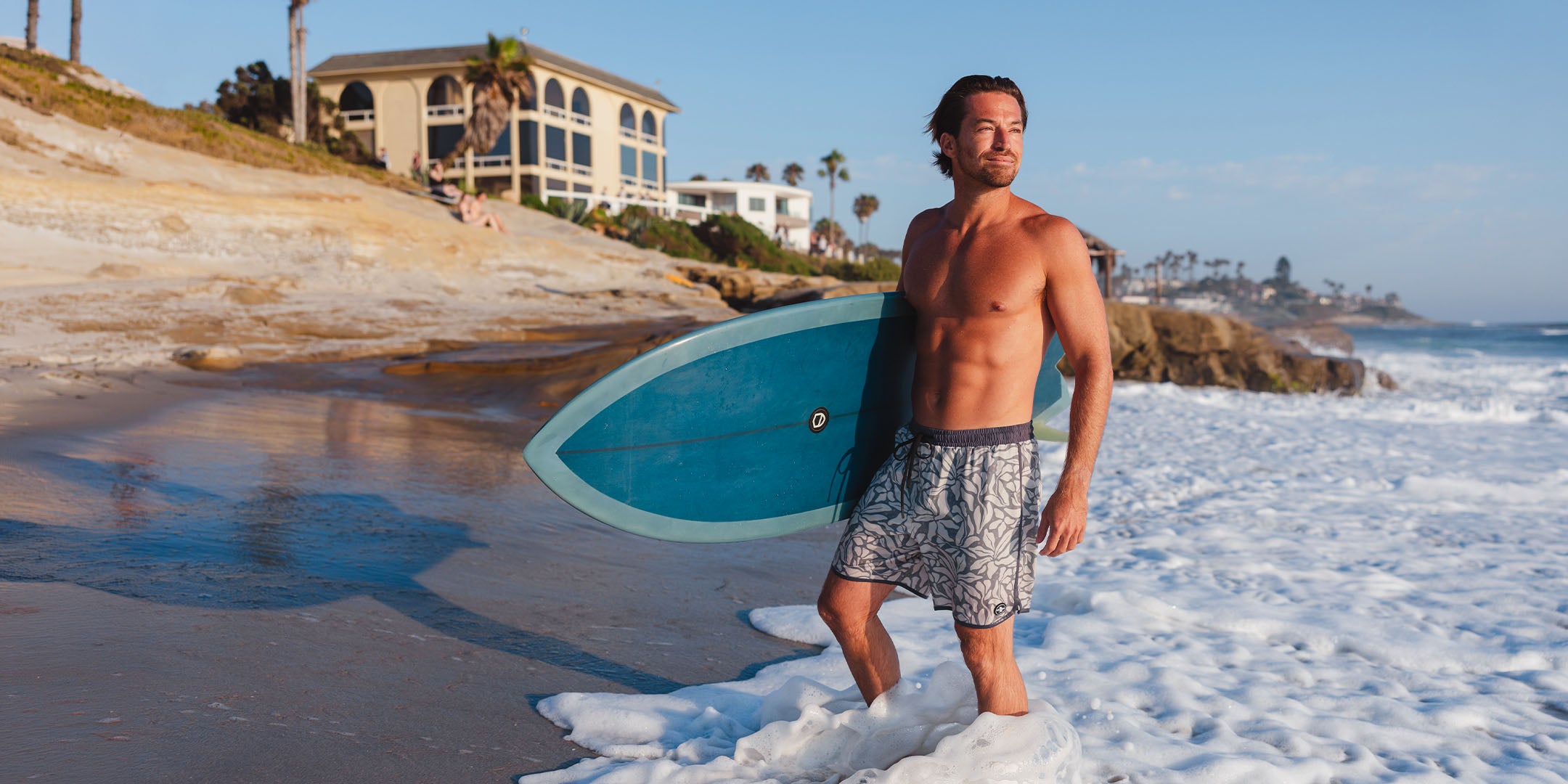 Man holding a surfboard on a beach with houses and palm trees in the background in La Jolla, California.