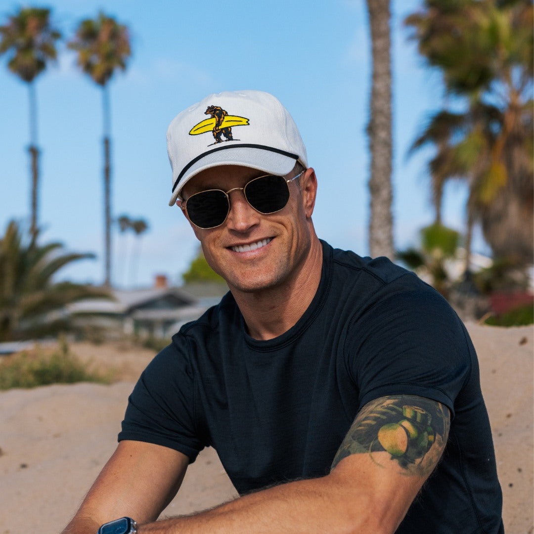 Man wearing a cap with bear holding a surfboard design, sunglasses, and black t-shirt on a beach with palm trees in the background.