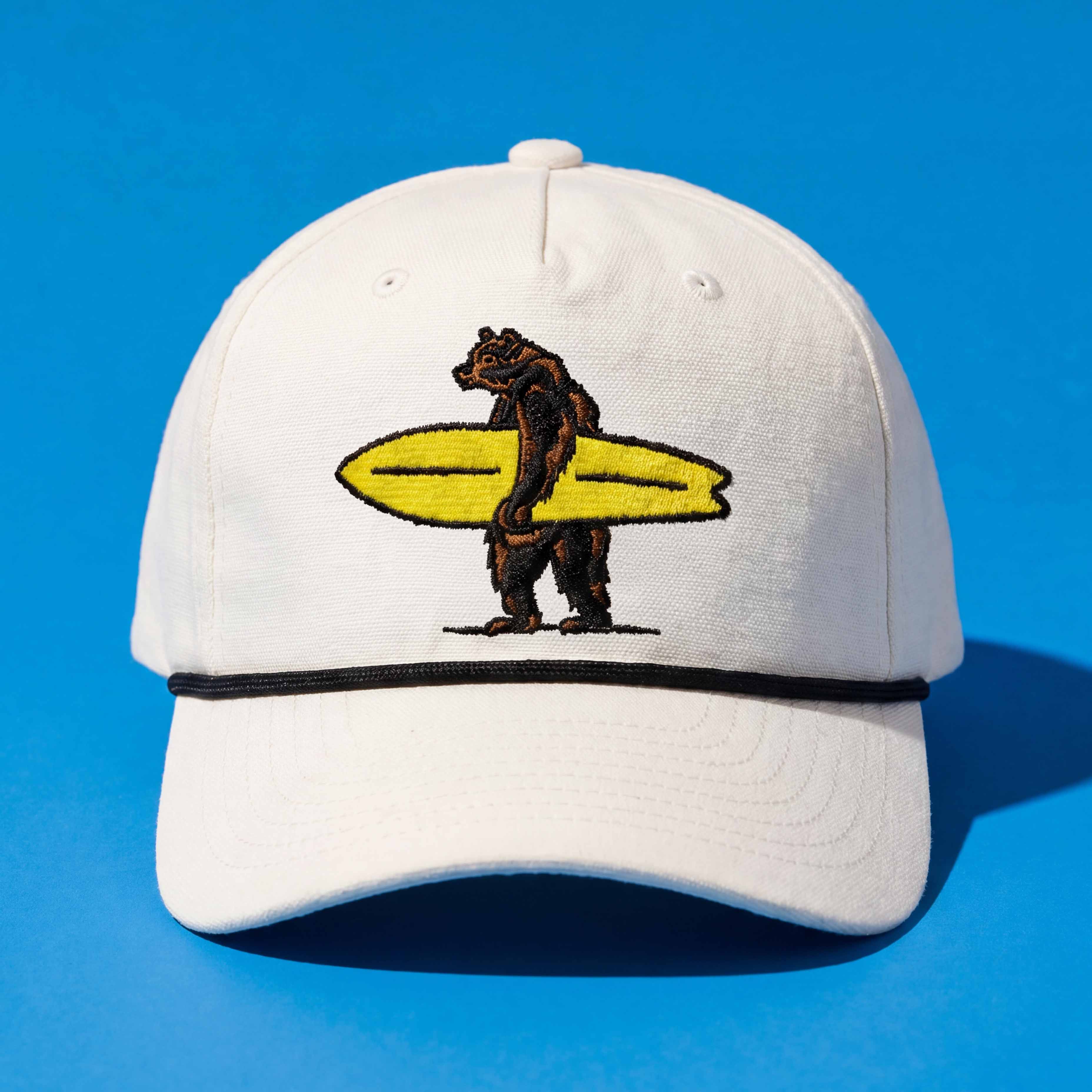 White cap with a bear holding a yellow surfboard design on a blue background