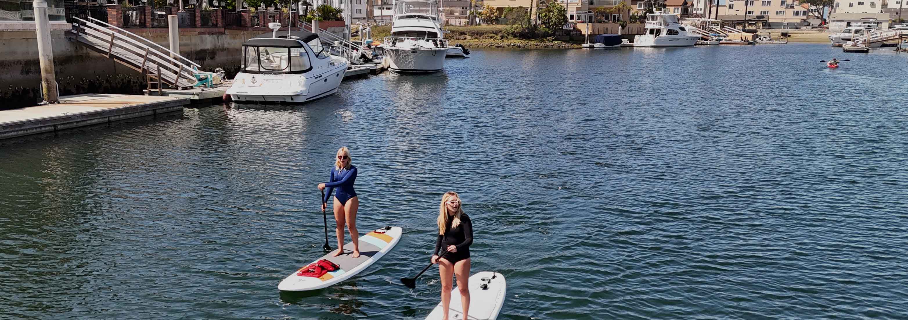 Two people paddleboarding on a calm body of water with boats in the background.