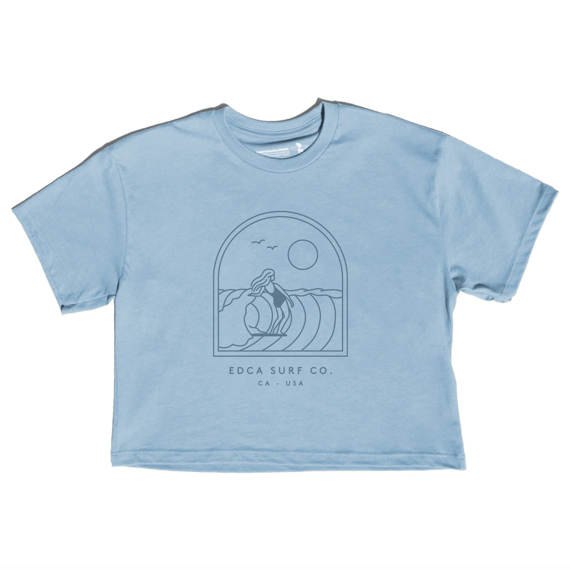 A light blue short-sleeve crop top t-shirt featuring a dark blue line art graphic of a surfer riding a wave inside an arch, with the text 'EDCA SURF CO.' and 'CA.