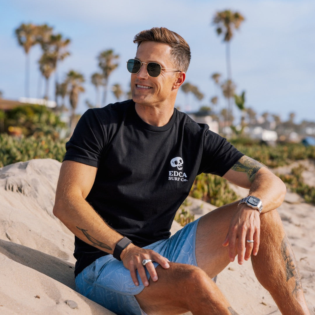 Man wearing sunglasses and a black t-shirt with 'EDCA Surf' logo sitting on sand with palm trees in the background