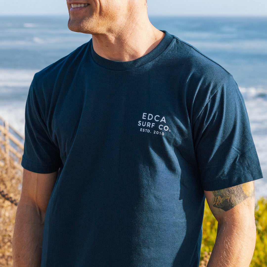 Man wearing a navy blue t-shirt with 'EDCA Surf Co.' logo by the ocean.