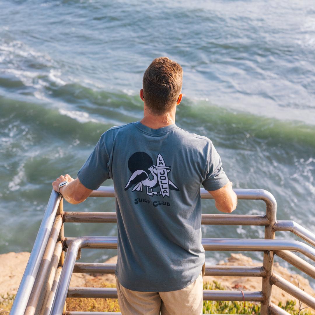 Man wearing a t-shirt with a pelican design with the text "La Jolla Surf Club", standing by a railing overlooking the ocean.