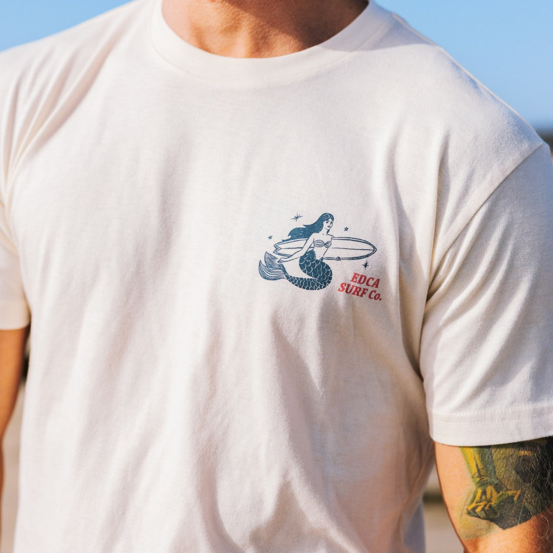 White t-shirt with mermaid graphic and 'EDCA Surf Co.' text on a sunny day.
