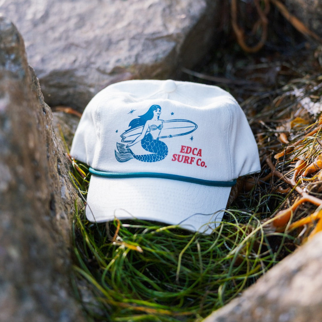 White cap with mermaid design and 'EDCA SURF CO.' text on grass and rocks