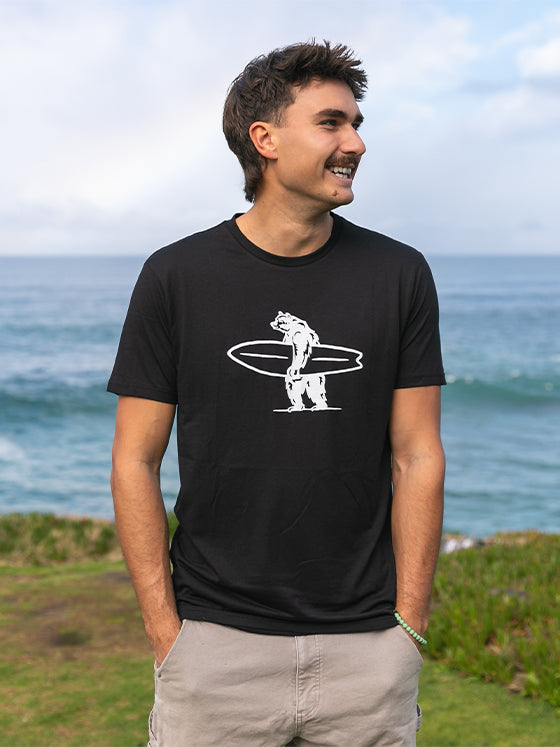 Man wearing a black t-shirt with a bear holding a surfboard graphic, standing outdoors with ocean in the background.