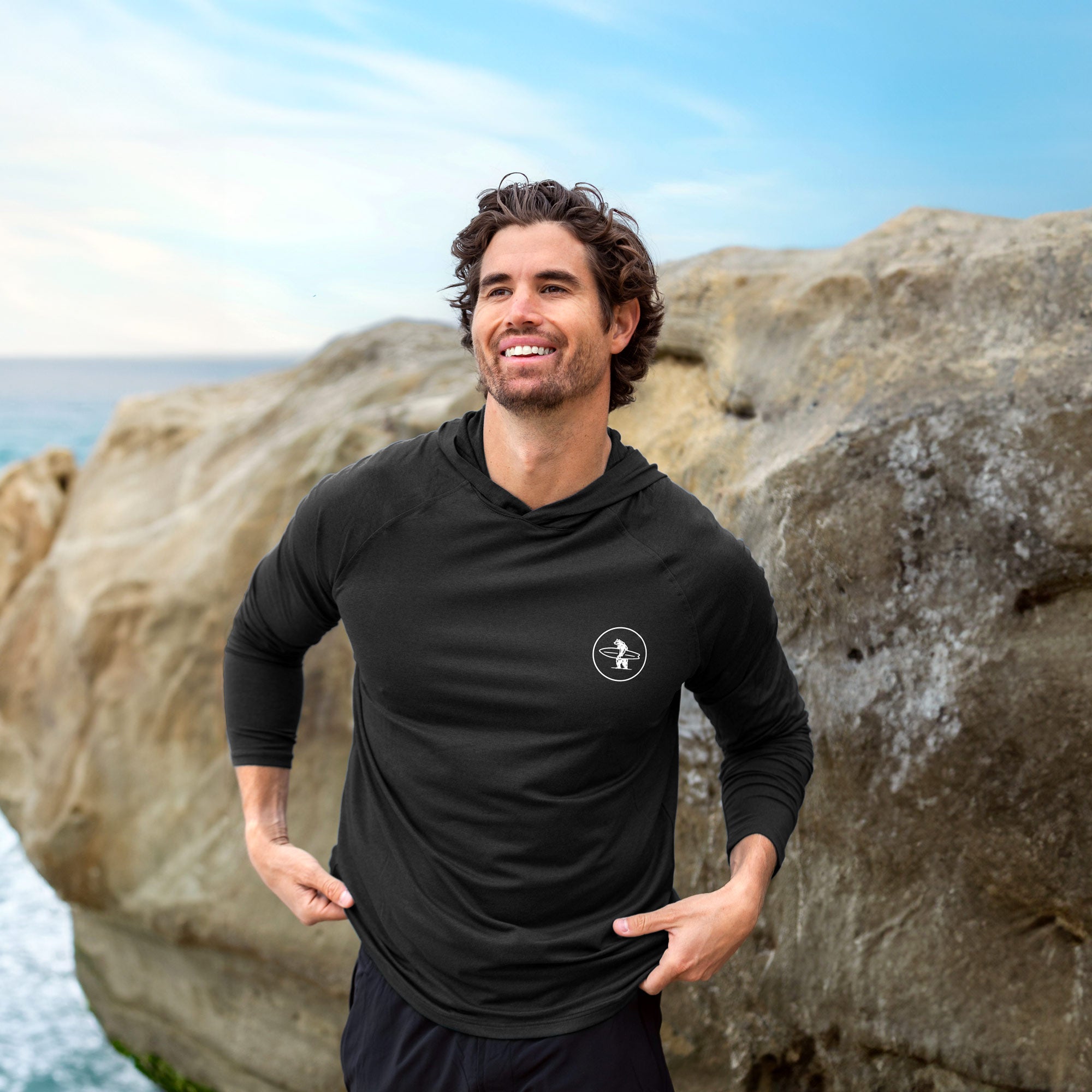 Male model at the beach wearing a black lightweight sun protective hoodie made out of performance fabric. The hoodie features a small white chest logo of a bear holding a surfboard. 