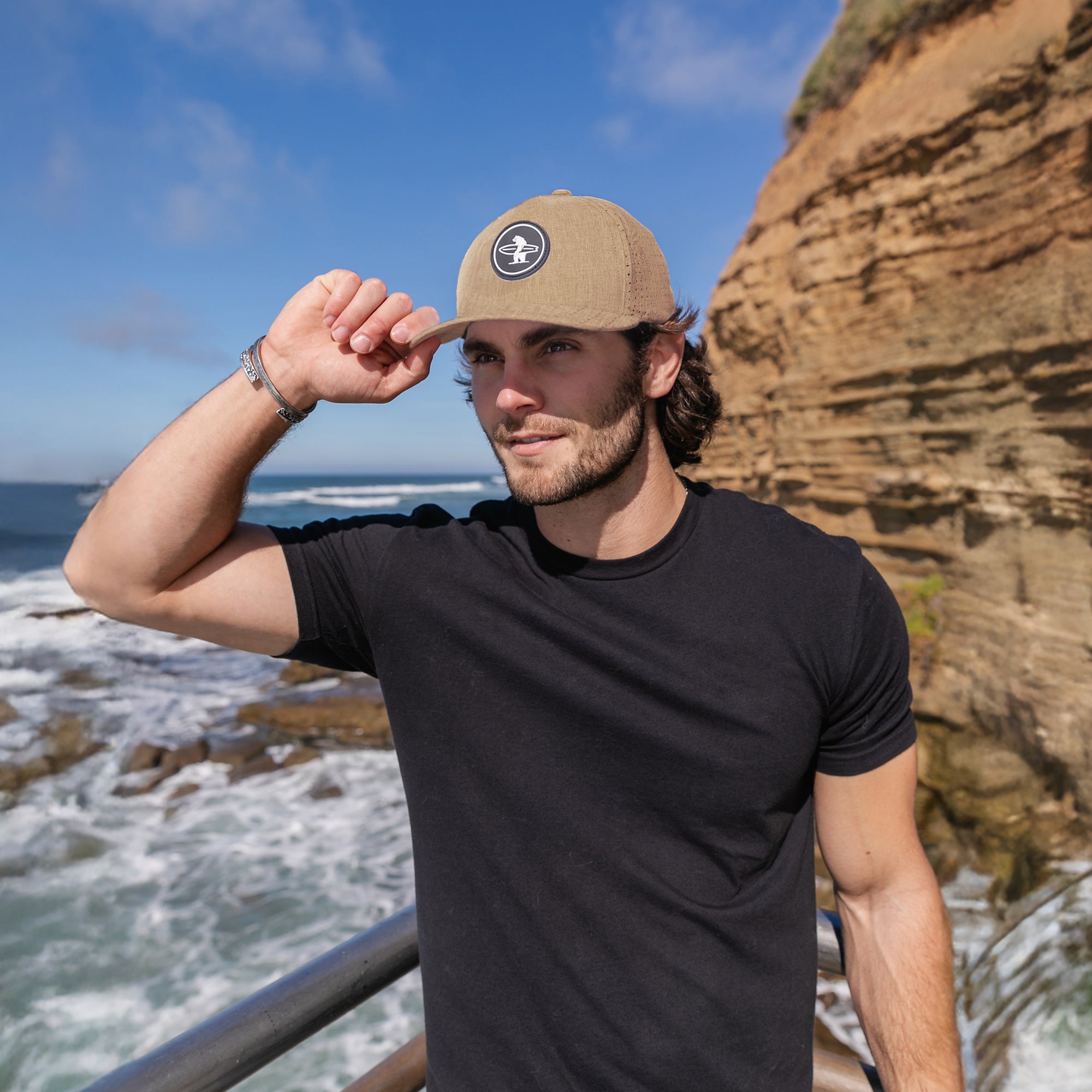 Man on the beach wearing a performance hat in the color light brown. The hat has a black rubber patch of a white bear holding a surfboard on it.