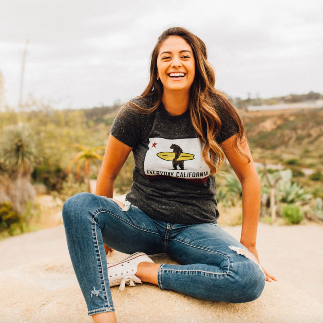 California girl sitting on a rock at the sandy beach wearing her Everyday California La Hermosa Tee