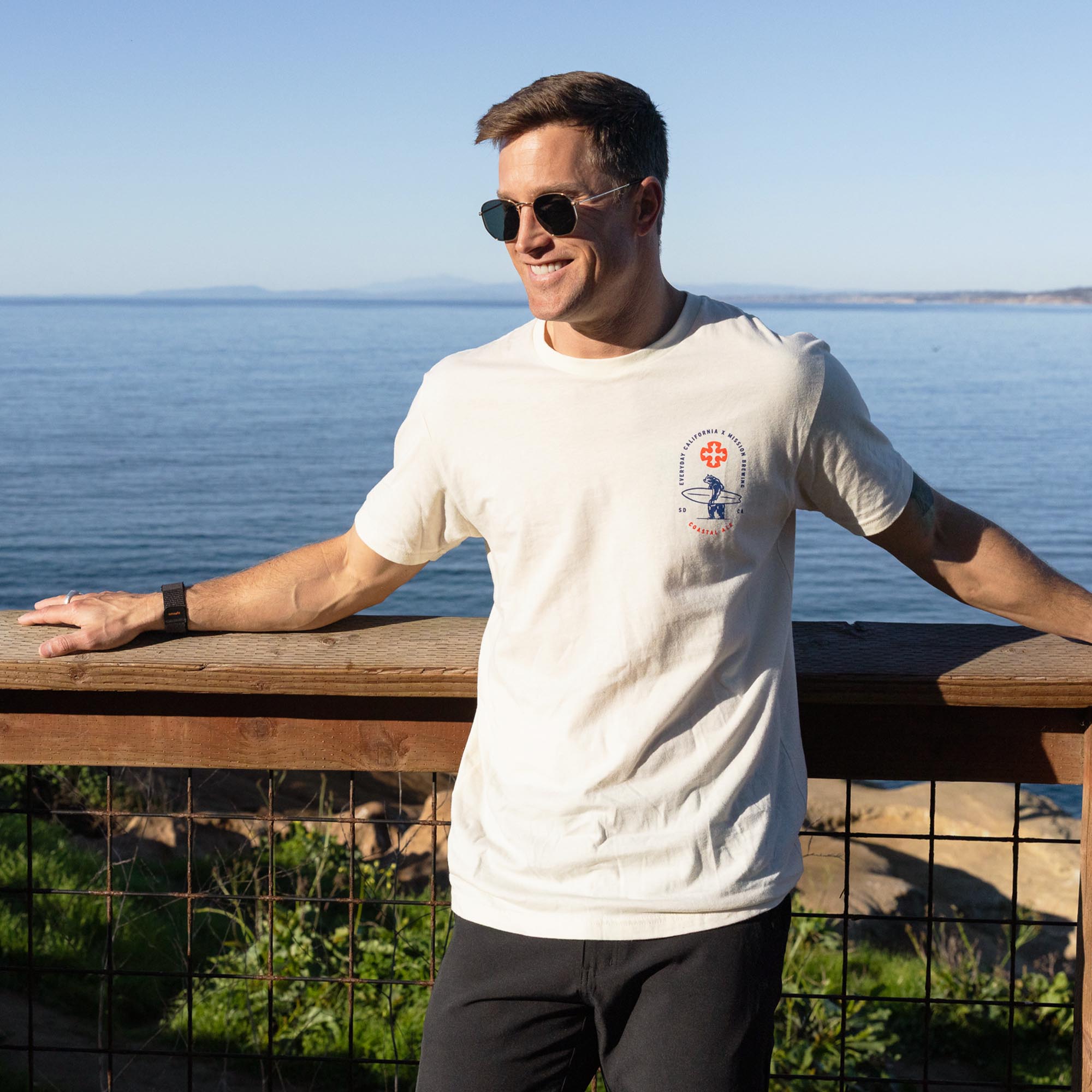 Man wearing a natural t-shirt with a logo, standing by a wooden railing with a scenic ocean view.