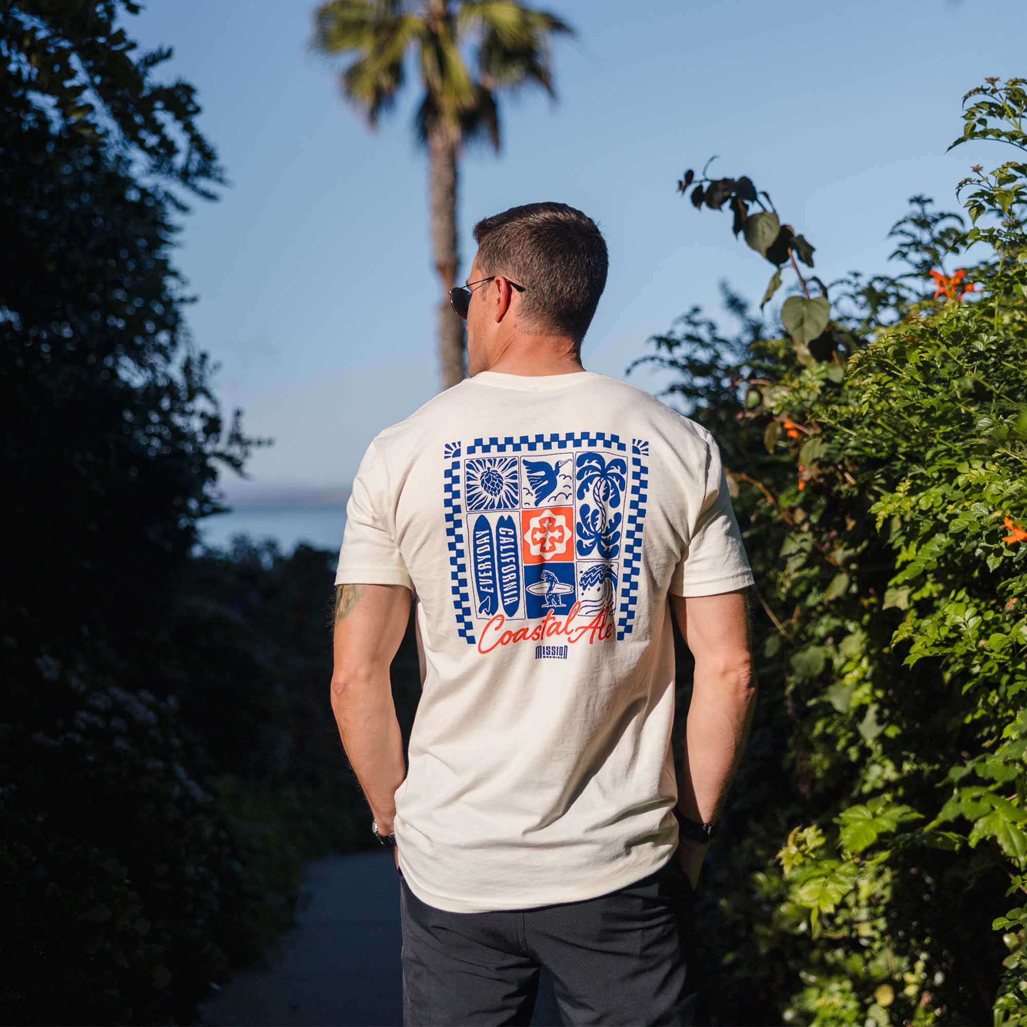 Man wearing a natural t-shirt with a coastal blue and red graphic design, standing outdoors with greenery around.