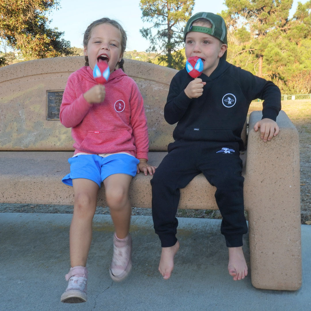 Two children sitting on a concrete bench outdoors, each holding a red and blue lollipop. The child on the left wears a pink hoodie and blue shorts, and the child on the right wears a black hoodie and black pants with a green cap.