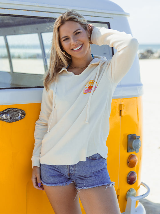 Girl smiling at the camera wearing a white hoodie standing in front of a bright yellow VW bus on the beach in San Diego, California. 