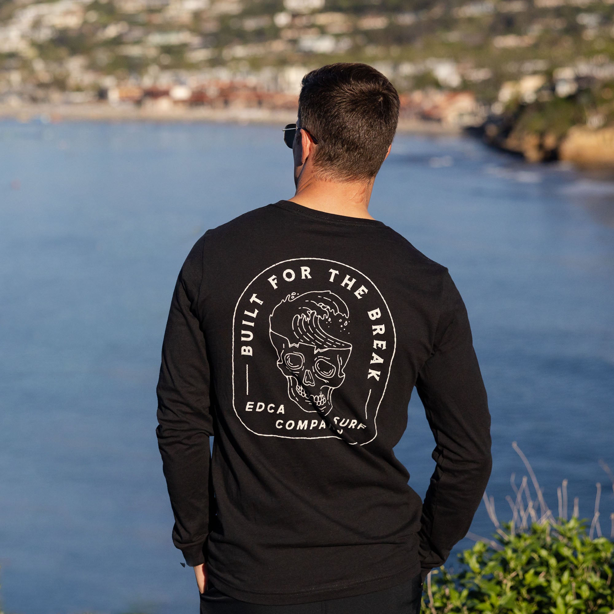 Male model wearing a black long-sleeve shirt with a graphic design, standing by a body of water.