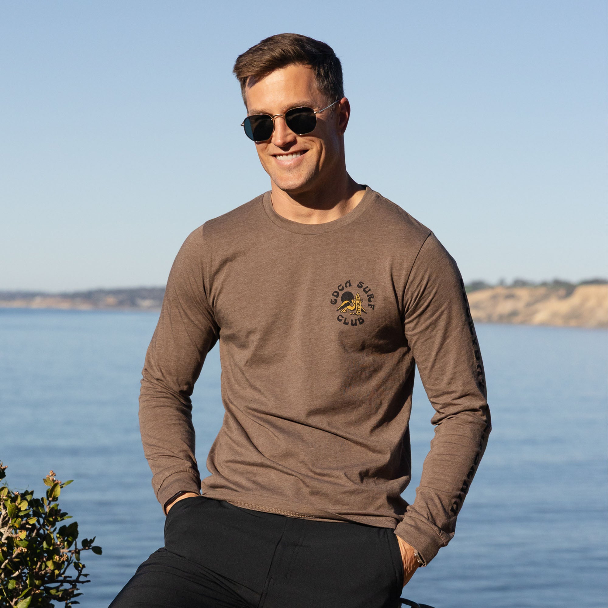 Man wearing an Everyday California brown long-sleeve shirt with a logo by a body of water